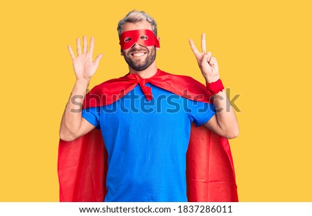 Young blond man wearing super hero custome showing and pointing up with fingers number seven while smiling confident and happy. 