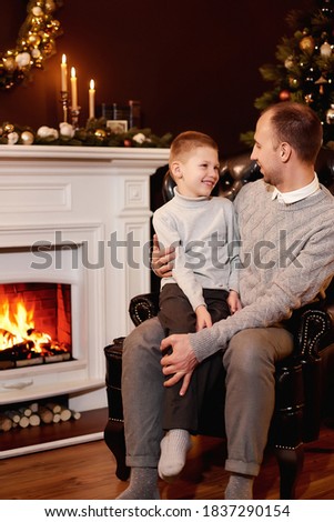 Father with his son sitting in armchair by Christmas tree and fireplace, looking at each other. Christmas wreath on the wall. On the fireplace there are candles and books,Xmas tree decorated by lights