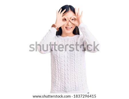 Young beautiful chinese woman wearing casual sweater doing ok gesture like binoculars sticking tongue out, eyes looking through fingers. crazy expression. 