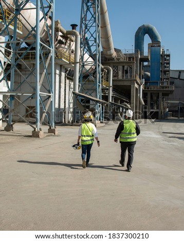 Workers of a cement plant, a man and a woman in helmets and specials are walking through the territory of the enterprise. Technological works for the production of cement.  Royalty-Free Stock Photo #1837300210
