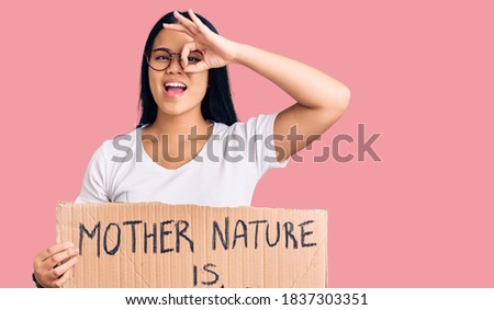Young beautiful asian girl holding mother nature is crying protest cardboard banner smiling happy doing ok sign with hand on eye looking through fingers 
