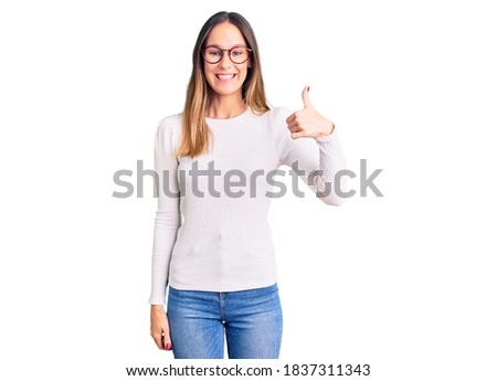 Beautiful brunette young woman wearing casual white sweater and glasses doing happy thumbs up gesture with hand. approving expression looking at the camera showing success. 