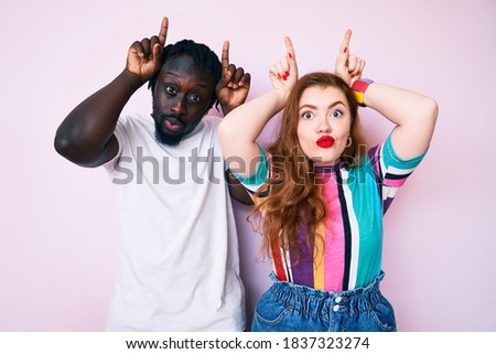 Interracial couple wearing casual clothes doing funny gesture with finger over head as bull horns 