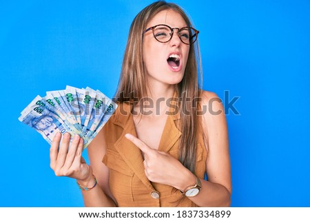 Young blonde girl holding south african rand banknotes angry and mad screaming frustrated and furious, shouting with anger. rage and aggressive concept. 