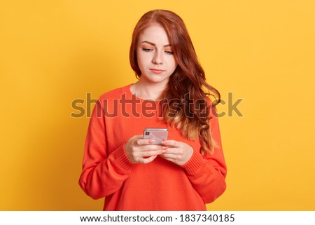 Serious young red haired woman girl in orange sweater, posing isolated on yellow wall, pensive girl using mobile phone, typing sms message or checking social networks.