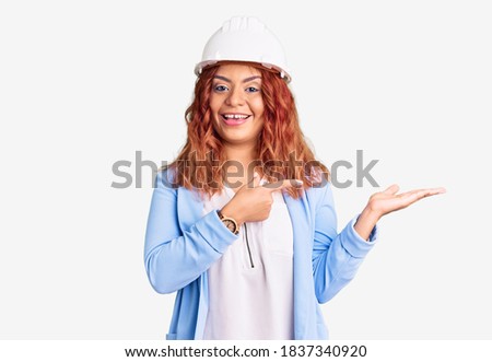 Young latin woman wearing architect hardhat amazed and smiling to the camera while presenting with hand and pointing with finger. 