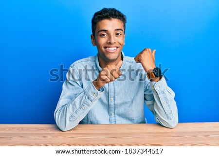 Young handsome african american man wearing casual clothes sitting on the table pointing to the back behind with hand and thumbs up, smiling confident 
