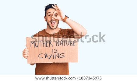 Young hispanic man holding mother nature is crying protest cardboard banner smiling happy doing ok sign with hand on eye looking through fingers 