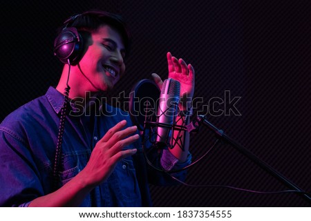 Young Asian man singing to condenser microphone in studio. Professional male vocalist wearing headphone while performing in voice recording room. Music production concept.