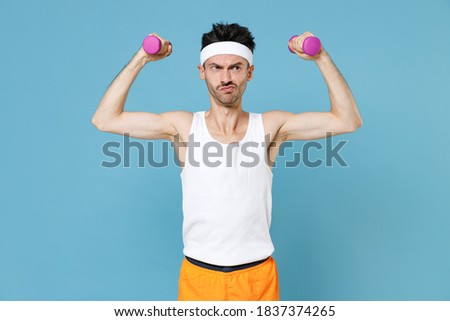 Confused young strong sporty fitness man with thin skinny body sportsman in white headband shirt shorts doing exercise with dumbbells isolated on blue background. Workout gym sport motivation concept Royalty-Free Stock Photo #1837374265