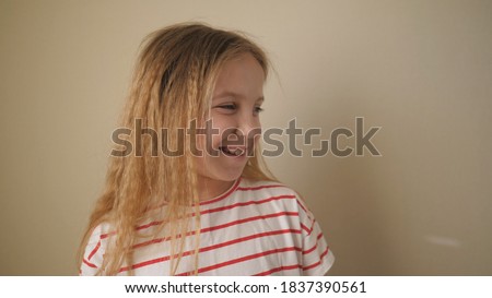 Little smiling female child looking to the side and laughing with someone indoor. Positive emotions of cute kid on her face. Happy small blonde girl against the background of wall. Close up Slow mo