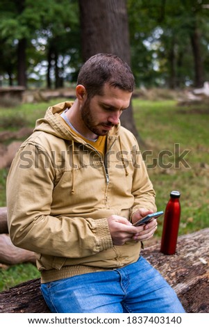 Photo of a young and attractive man taking a break from a excursion in the nature sitting on a tree resting and checking his phone. Surrounded my trees in the forest.