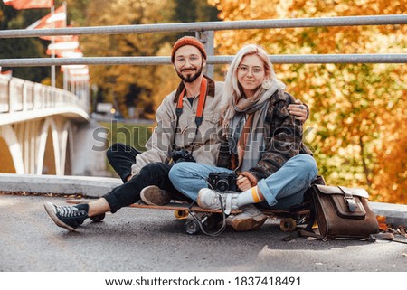 Bearded guy and blond girl relaxing embracing in nature sitting on skate in autumn landscape background.
