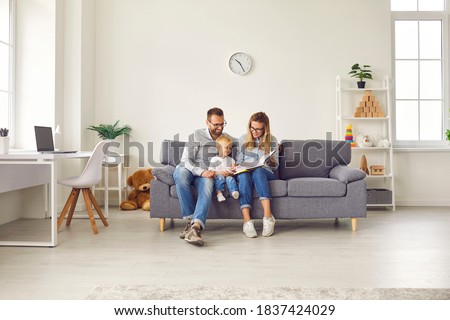 Happy young family with child sitting on comfy soft gray couch in spacious studio apartment or living-room of new house, reading book together, enjoying quiet leisure time on weekend at home Royalty-Free Stock Photo #1837424029