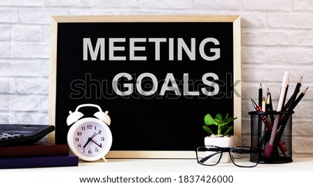 On the desktop against the background of a light brick wall in a stand are pencils, a white alarm clock and a black board with the inscription MEETING GOALS.