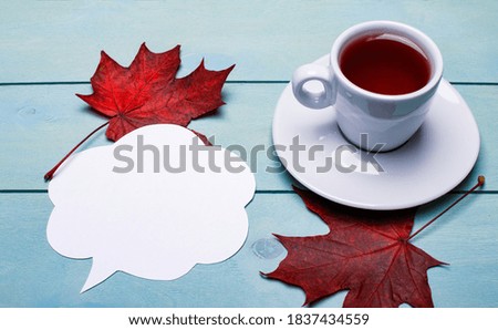 On a blue wooden background, burgundy maple leaves, a white cup with tea and a white sheet for writing. Copy space. Autumn concept