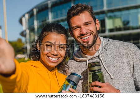 20s mixed race happy couple taking selfie after jogging holding bottles of water in urban area