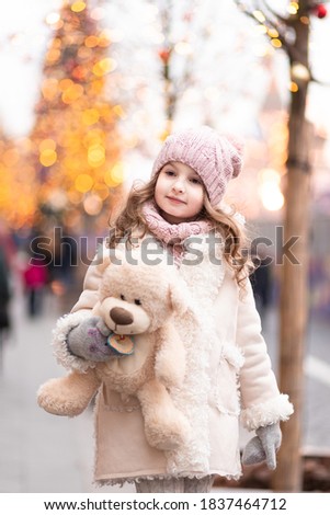 A beautiful winter girl in a pink hat and a sheepskin coat, holding a soft toy bear at the New Year's fair against the background of a Christmas tree. Christmas holidays concept. Selective focus