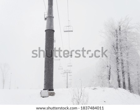Empty chair lift in fog on winter day during snowfall on background of trees