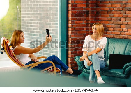 Happy and relaxed female friends sitting on the sofa and rocking chair, in the living room. One takes a selfie while rocking in a chair and the other woman is watching cute. Big city life