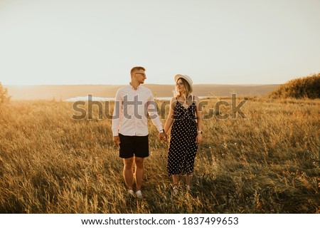 a guy with a girl in hat walking in the meadow. A couple of fair-haired fair-skinned people in love are resting in nature in a field at sunset.