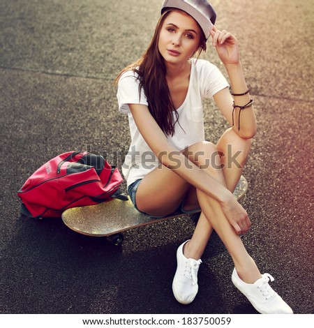 Fashion outdoor summer portrait of pretty brunette girl sitting on the ground with skateboard and red bag in hat. Summer colorful picture