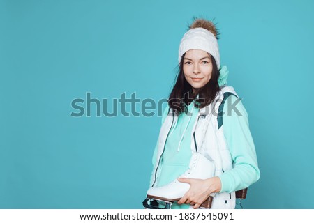 Winter hobby, ice skating. A brunette woman in a hoodie with figure skating skates on her shoulder. Studio shot on a blue background