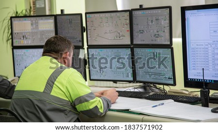 A male engineer, in special clothing, sits against the background of monitors depicting process monitoring. Technological works for the production of cement. Working atmosphere with copy space. Royalty-Free Stock Photo #1837571902