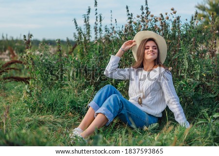Young beautiful brunette woman dressed in a white sweater, jeans and cowboy straw hat sitting on green grass, smiling and laughing during sunset