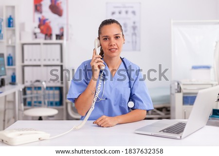 Portrait of medical nurse smiling at camera while using phone in hospital office. Health care physician sitting at desk using computer in modern clinic looking at monitor.
