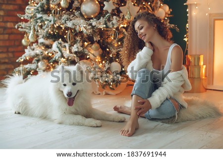 Beautiful woman sits on a white blanket with a dog hugs, against the background of a Christmas tree fireplace with candles in a decorated room. Happy New Year and Merry Christmas. High quality photo.