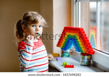 Cute little toddler girl by window create rainbow with colorful plastic blocks during pandemic coronavirus quarantine. Children made and paint rainbows around the world as sign. Kid eating lollipop.