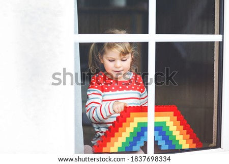 Cute little toddler girl by window create rainbow with colorful plastic blocks during pandemic coronavirus quarantine. Children made and paint rainbows around the world as sign.