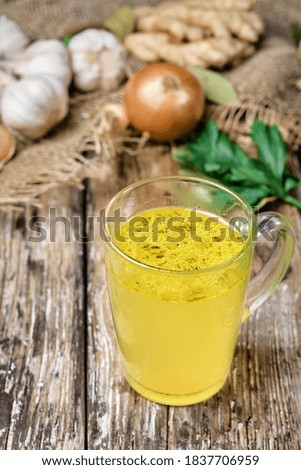 Glass mug with chicken broth on a dark wooden table. Close-up. Ingredients for chicken soup in the background