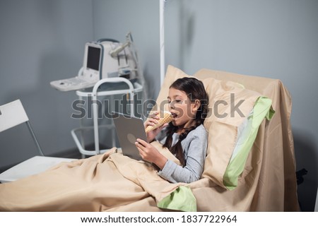 Little brunette girl eating an ice cream and watching cartoons on her tablet in a hospital bed.