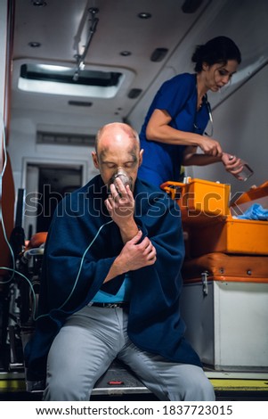 Injured man in a blanket breathing through an oxygen mask in an ambulance car, nurse searching for something in her medical kit in the background.