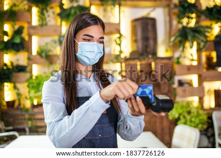 Young waitress with protective face mask making a credit card payment with card reader machine. Credit card swipe through terminal in restaurant. Waitress hand swiping debit card to pay the bill