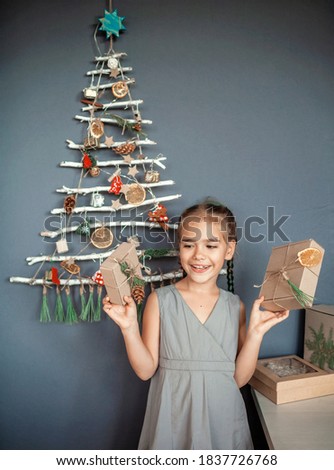 Happy girl holding present box near original alternative Christmas tree made with frozen branches and decorated with tiny ecological toys and paper garland, zero waste and plastic free holidays