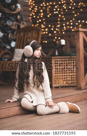 Stylish kid girl 5-6 year old wearing knitted clothes and fluffy headphones sitting outdoors over Christmas lights close up. New Year. Winter holiday season. 