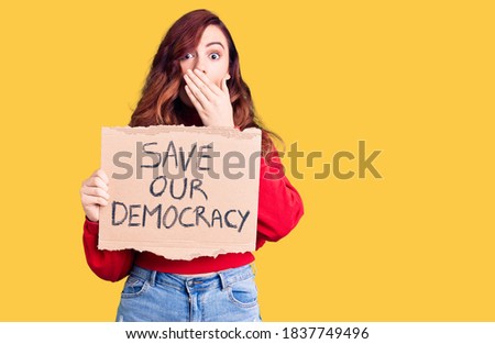 Young beautiful woman holding save our democracy protest banner covering mouth with hand, shocked and afraid for mistake. surprised expression 