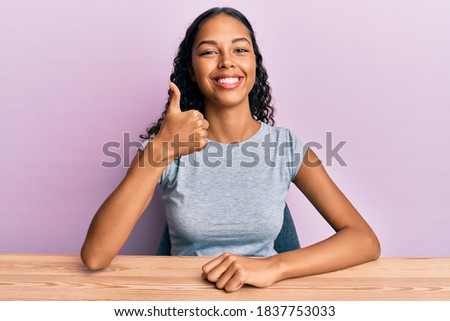 Young african american girl wearing casual clothes sitting on the table doing happy thumbs up gesture with hand. approving expression looking at the camera showing success. 