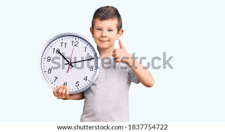 Cute blond kid holding big clock smiling happy and positive, thumb up doing excellent and approval sign 