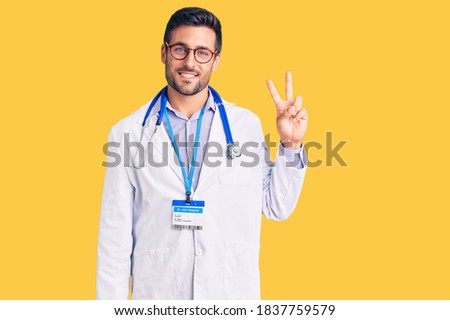 Young hispanic man wearing doctor uniform and stethoscope smiling looking to the camera showing fingers doing victory sign. number two. 