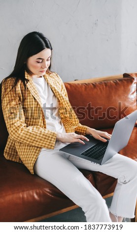 Portrait of focused beautiful woman sitting on a sofa, looking at laptop screen on video call or chat and typing on keyboard. Businesswoman working distantly at home office.