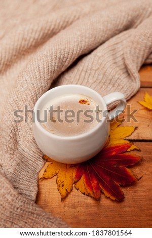 vertical cozy picture of Breakfast, coffee with milk foam in a white mug, autumn red and yellow maple leaves and a knitted beige sweater on a wooden table,selective focus
