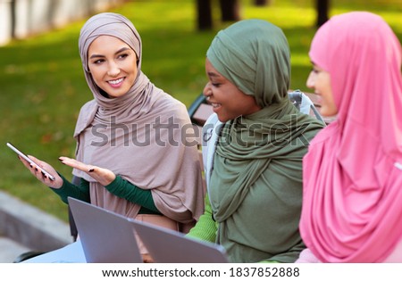 Modern Muslim Student Lady Recommending Smartphone Mobile Application To Friends Sitting On Bench Learning With Laptop Computers In Park Outdoors. Educational Application For Phone. Selective Focus