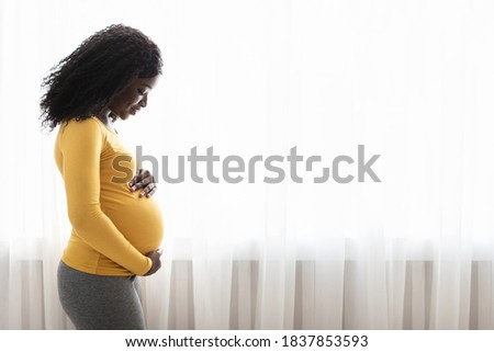 Beautiful pregnant black woman hugging her tummy, enjoying her pregnancy, free space. Side view of african american expecting lady standing next to window at home, touching her big belly Royalty-Free Stock Photo #1837853593
