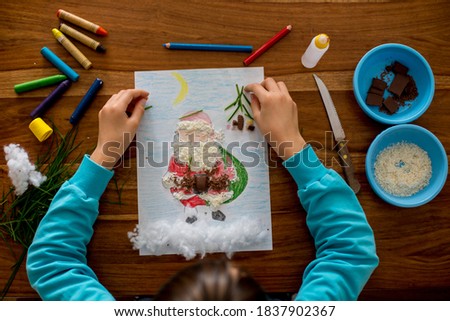 School child, boy, drawing a picture of Santa Claus and decorating him with different elements - chocolate, coconut, grass and cotton