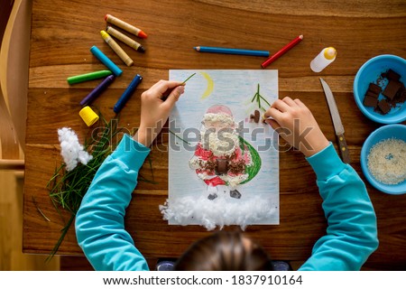 School child, boy, drawing a picture of Santa Claus and decorating him with different elements - chocolate, coconut, grass and cotton