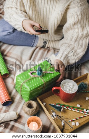 Female hand with smartphone takes pictures of New Year's or Christmas flatlay for social networks. Creating content. Wooden tray with cocoa mug marshmallows, toy tree, candle, stars, wrapped gift.